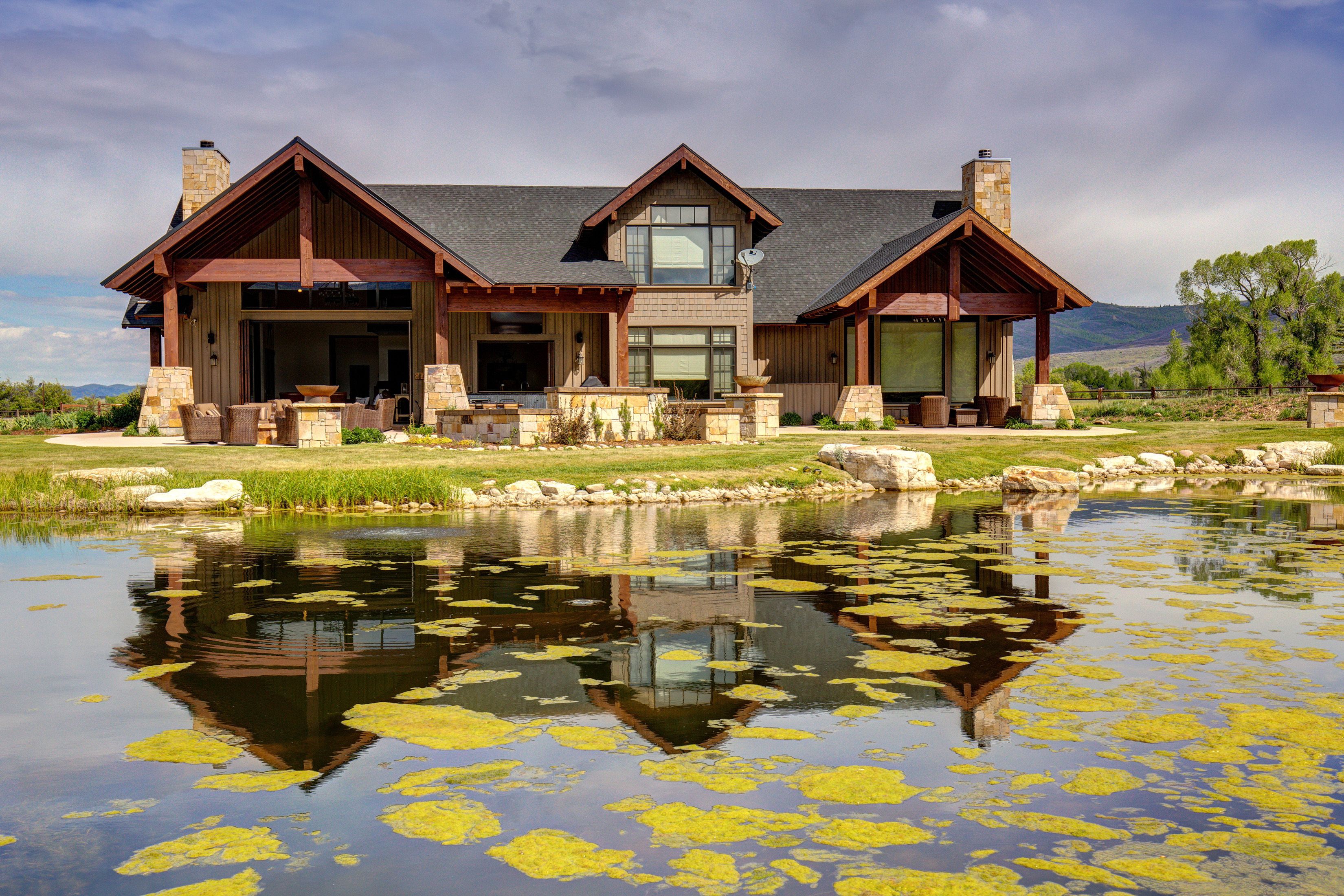 A large, two-story house with a stone chimney, a large window, and a pond in the foreground.