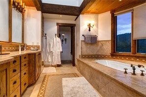 A spacious bathroom with a large bathtub, wooden cabinetry, and a view of the mountains through the window. - Abode on Jordanelle Way (Park City)