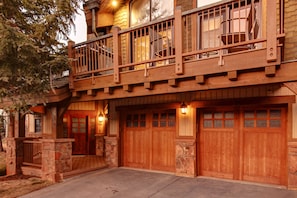 A two-story log cabin with a balcony, stone foundation, and wooden garage doors.