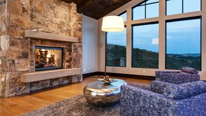 Ein Kamin aus Stein mit integriertem Fernseher, ein großes Fenster mit Blick ins Freie und eine moderne Sitzecke mit einer strukturierten Couch und Hocker.