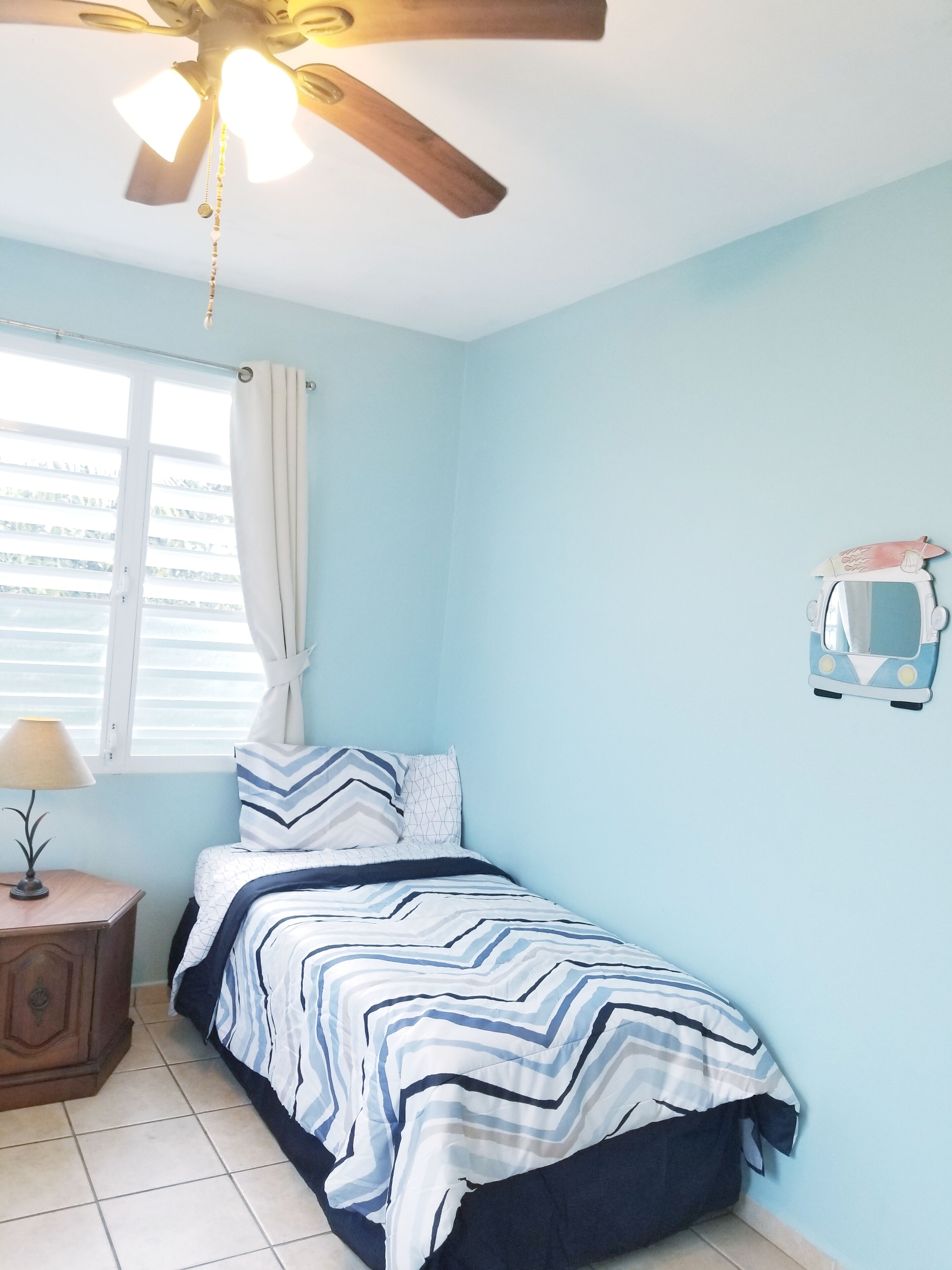 A bedroom with a bed, a nightstand, a ceiling fan, a window with curtains, and a wall-mounted decoration.