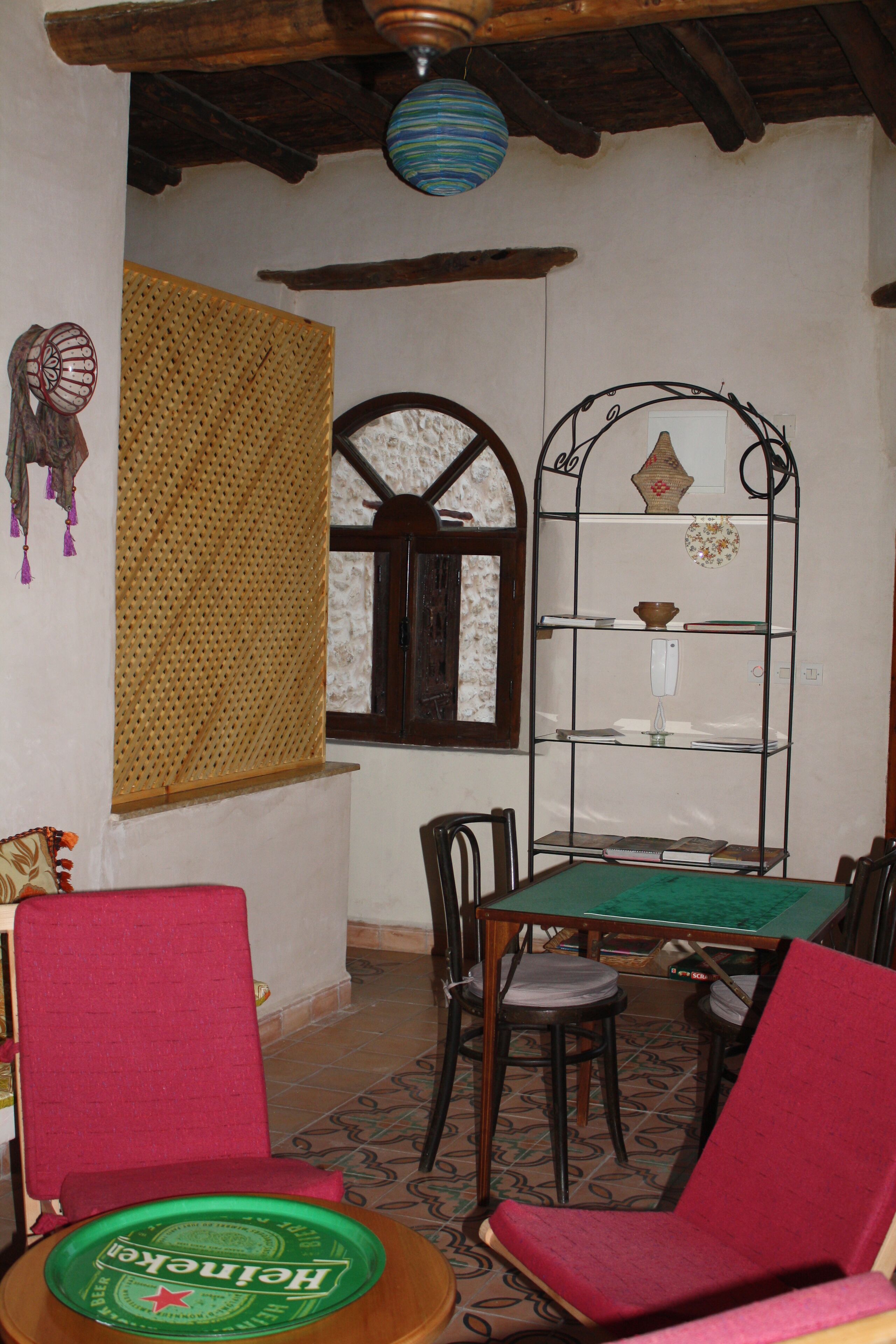 A room with a wooden ceiling, a window with a decorative frame, a metal shelf with books, a table with chairs, and a green tablecloth.