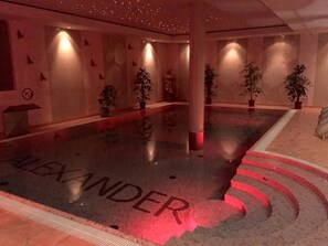 Ein Schwimmbecken mit dem Namen 'ALEXANDER' auf dem Boden, umgeben von Wänden mit einem Muster aus kleinen, dunklen Formen.
