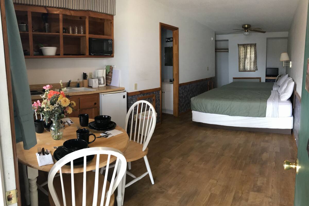 A compact hotel room with a dining area, a bed, a ceiling fan, and a small kitchenette.