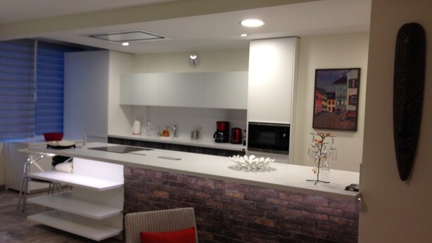 A modern kitchen with white cabinets, a brick accent wall, a dining area with a white table and chairs, and a cityscape painting on the wall.