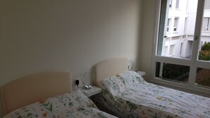 A bedroom with two single beds, a bedside table with a lamp, and a window with a view of buildings.