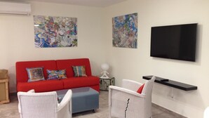 A living room with a red sofa, two white armchairs, a blue ottoman, a flat-screen TV, and two abstract paintings on the walls.