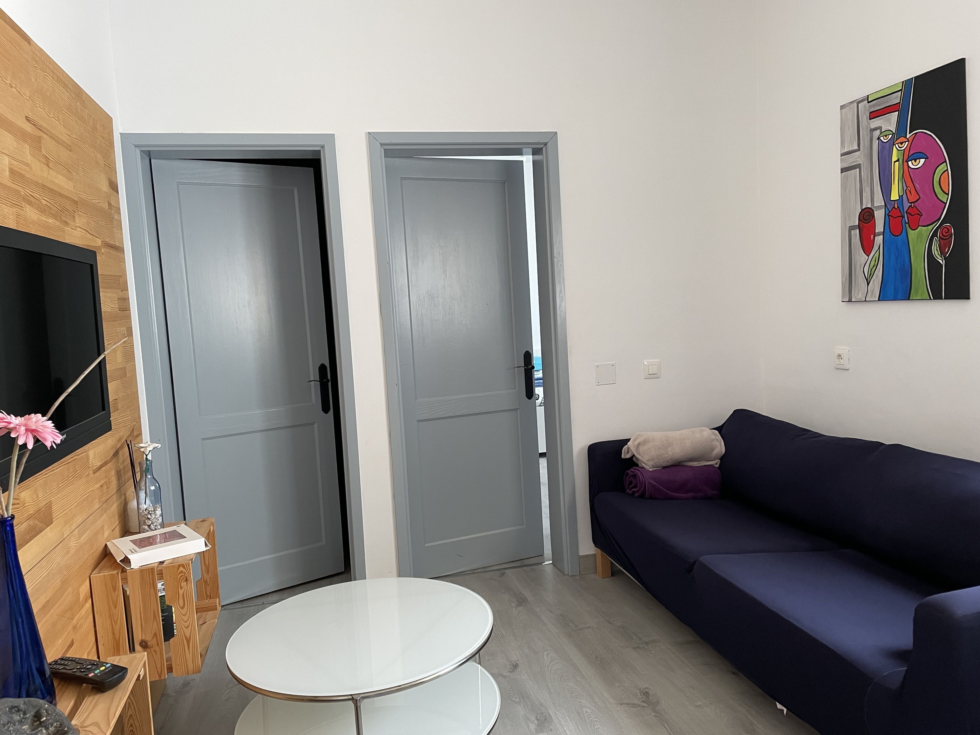 Una sala de estar moderna con un sofá azul oscuro, una mesa de centro blanca y un televisor montado en una pared de madera. Se observan dos puertas grises y un cuadro abstracto colorido en la pared.