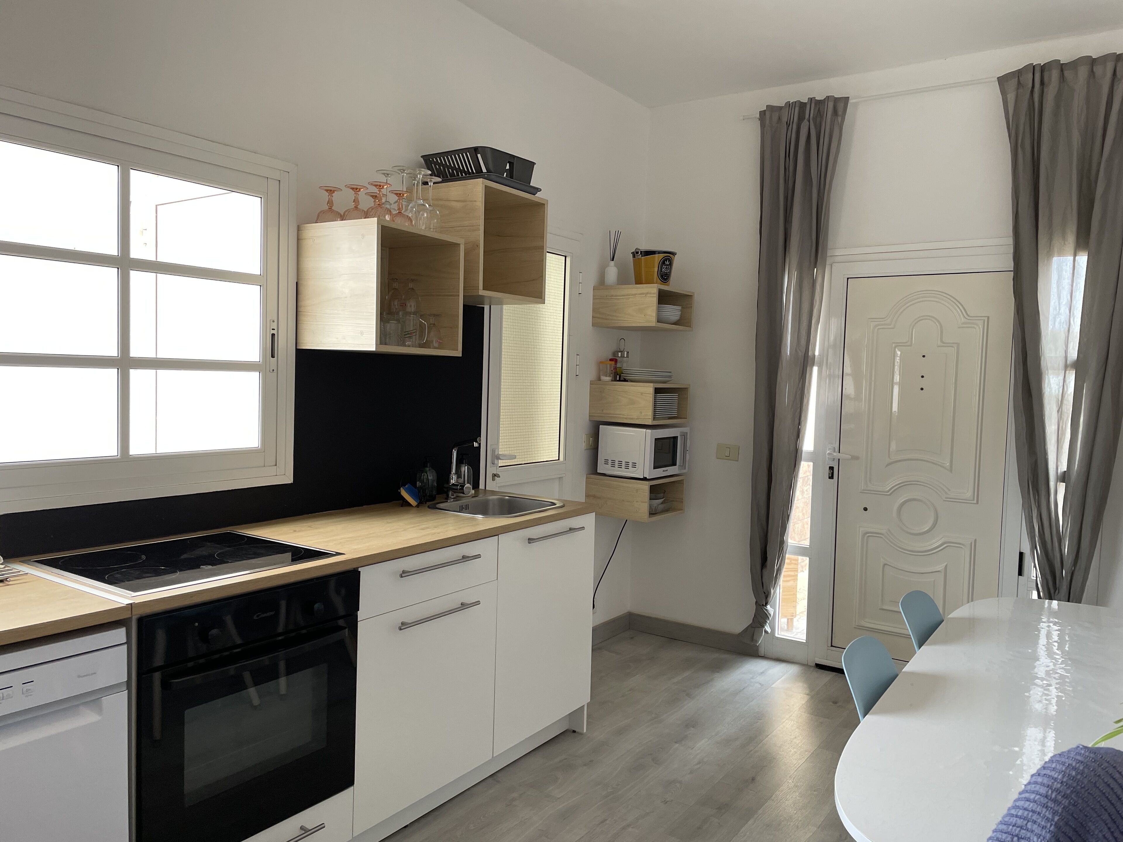 A modern kitchen with a built-in oven, a sink, and wooden cabinets. There is a dining table with chairs and a window with curtains.
