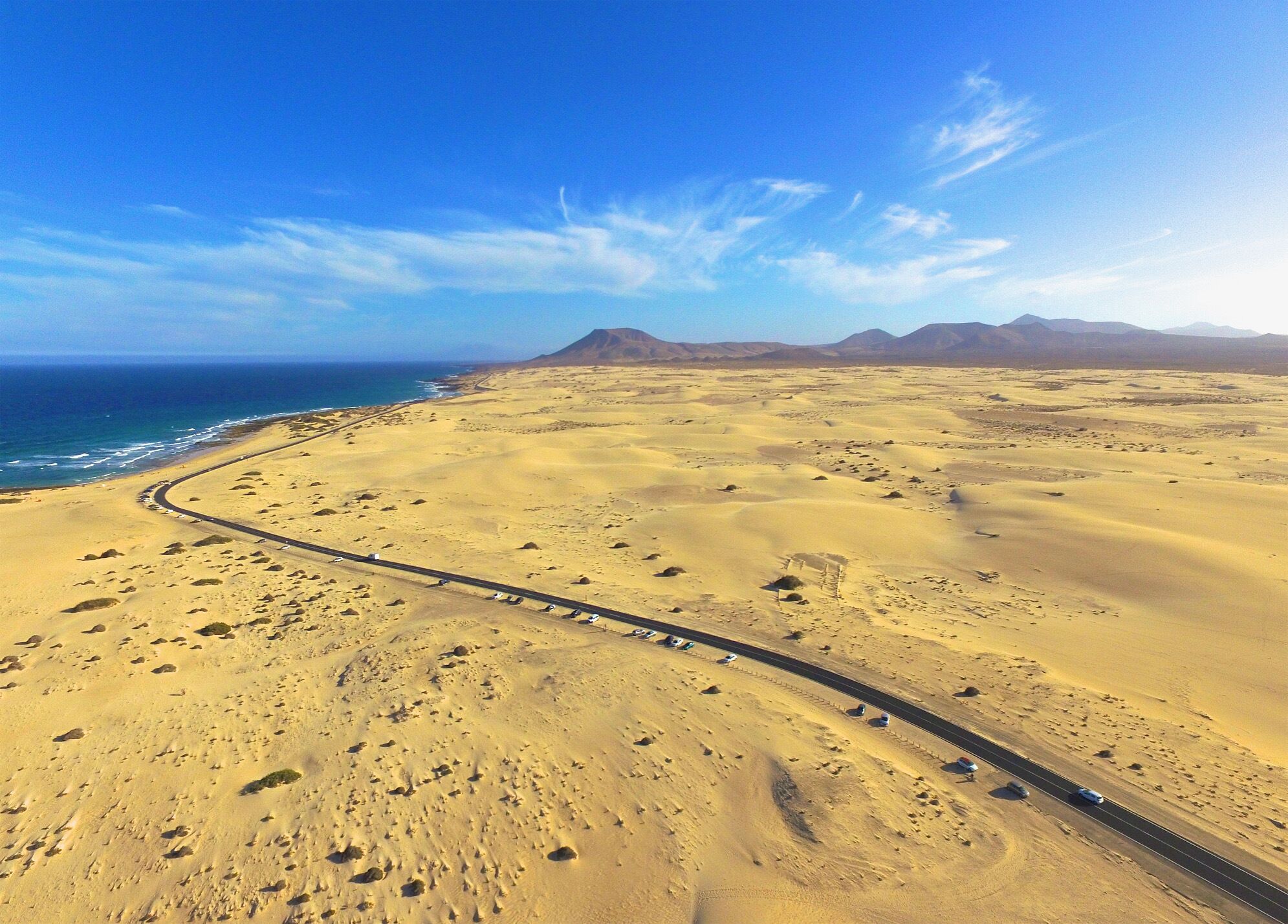 Un paysage désertique traversé par une route sinueuse qui mène à une plage et à des montagnes à l’horizon.