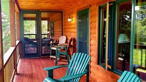 A wooden porch with a view of the forest, a table with chairs, and a grill.