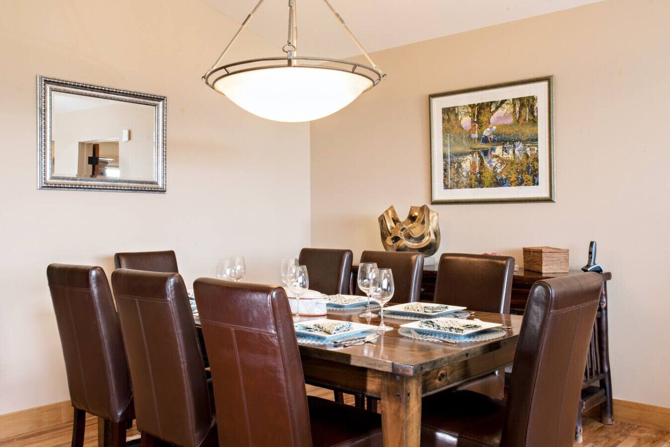 A dining room with a wooden table, brown leather chairs, and a chandelier. There is a framed painting on the wall and a mirror above the table.