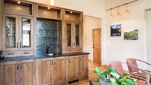 Una cocina moderna con armarios de madera, una encimera negra y un lavamanos. Hay una zona de comedor con una silla roja y una planta.