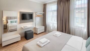 A modern hotel room with a large bed, a flat-screen TV mounted on the wall, a bedside table with a lamp, a small seating area, and a view of the outside through sheer curtains.