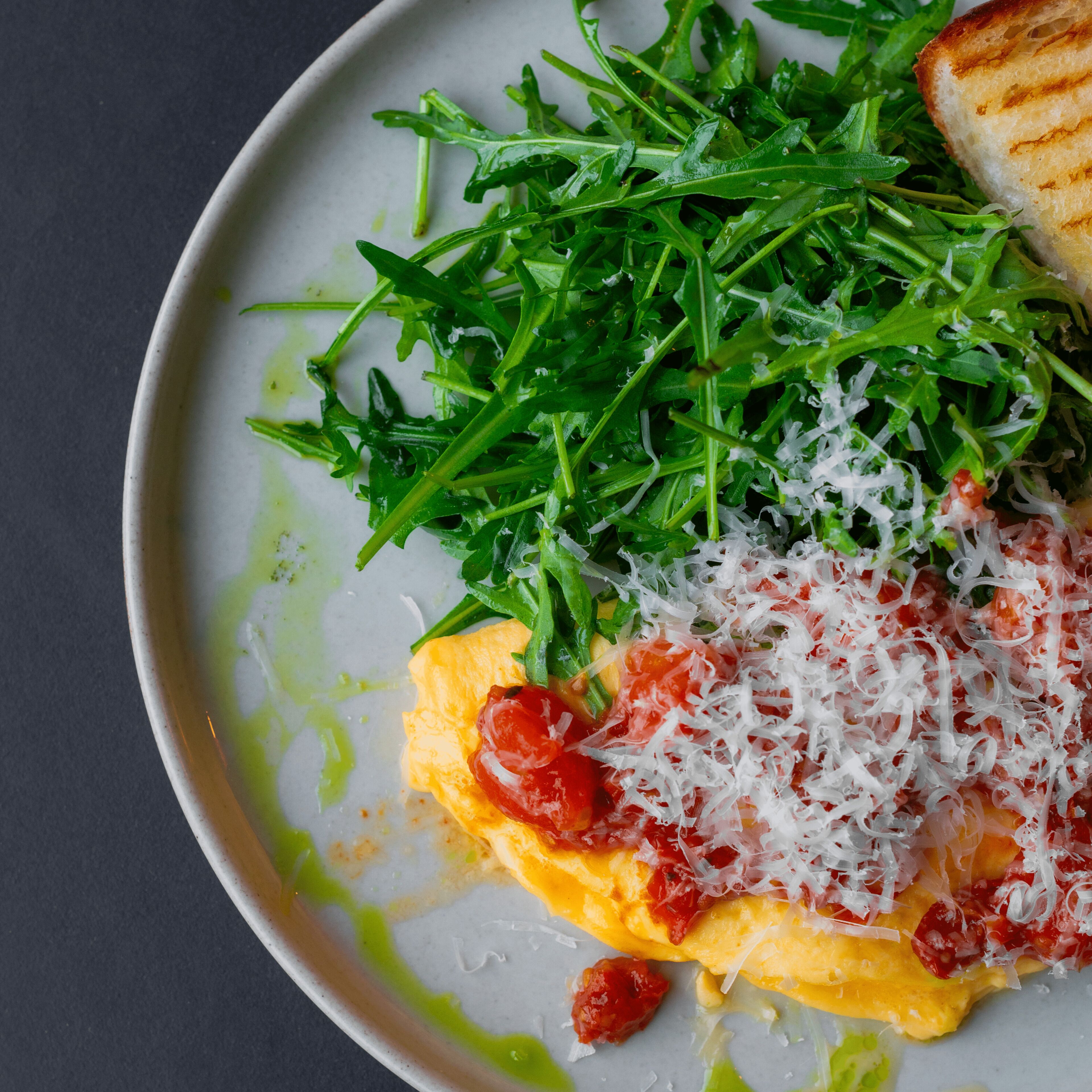 Ein Teller mit Rucola, gehobeltem Käse und einem gegrillten Gericht, möglicherweise einem Sandwich oder Fladenbrot, auf weißem Geschirr.