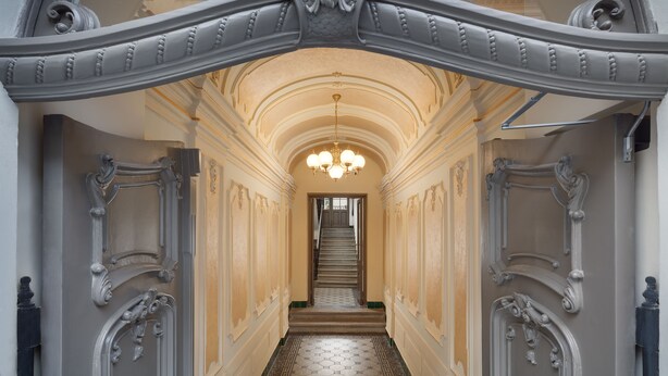 A grand hallway with ornate white railings, a central staircase, and a chandelier. - Louren Apartments (Prague)