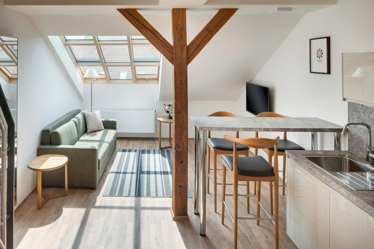 A modern loft apartment with a kitchen, dining area, and living space. It features a skylight, wooden beams, and a large window.