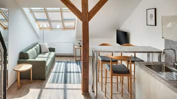 A modern loft apartment with a kitchen, dining area, and living space. It features a skylight, wooden beams, and a large window.