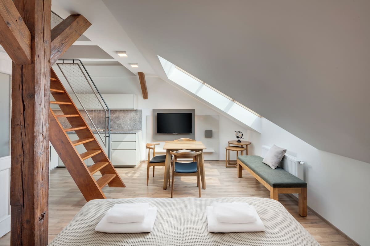 A modern loft bedroom with a sloped ceiling, a wooden staircase, a dining area, and a large TV.