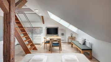 A modern loft bedroom with a sloped ceiling, a wooden staircase, a dining area, and a large TV.