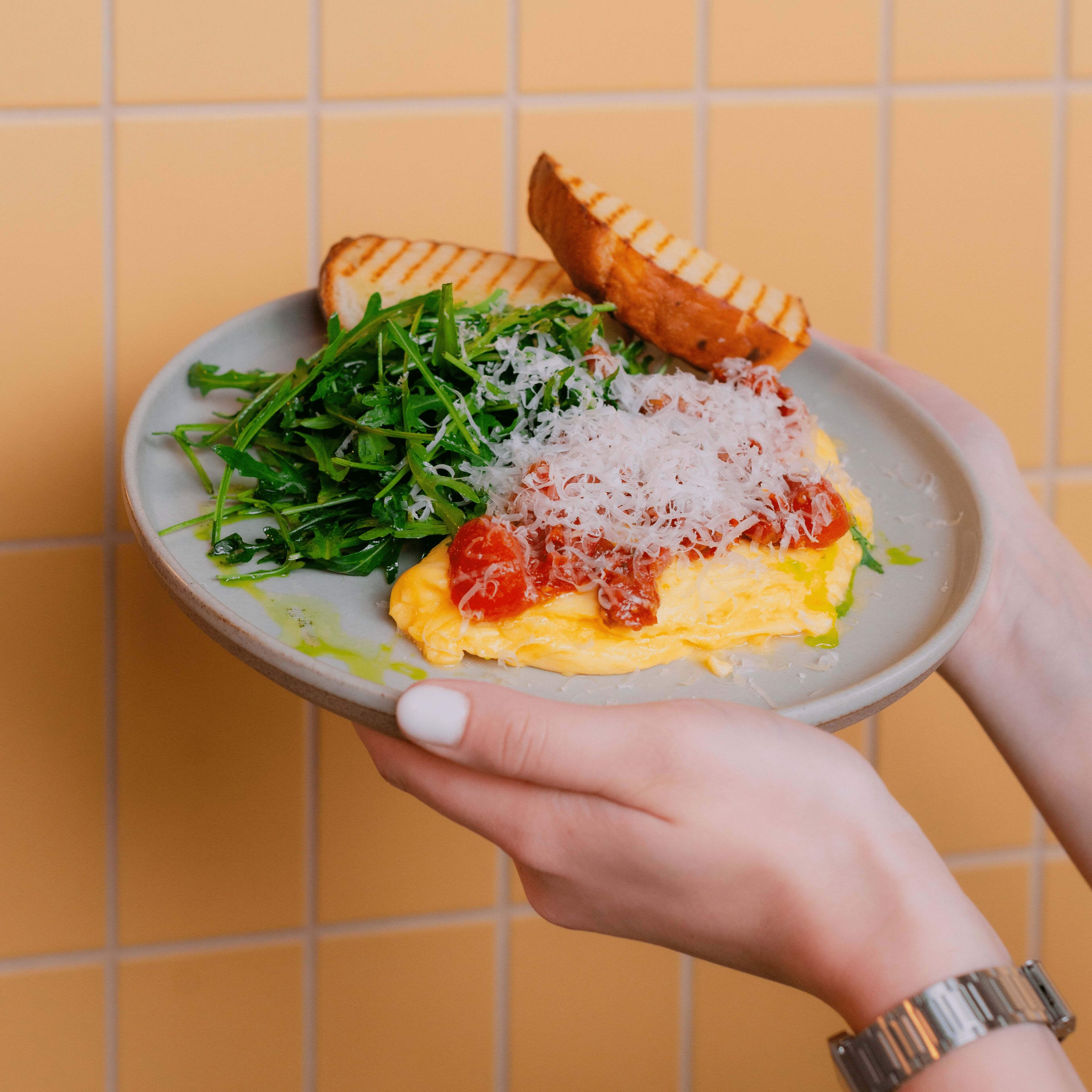 A plate with scrambled eggs, arugula, grated cheese, and a grilled bread slice.