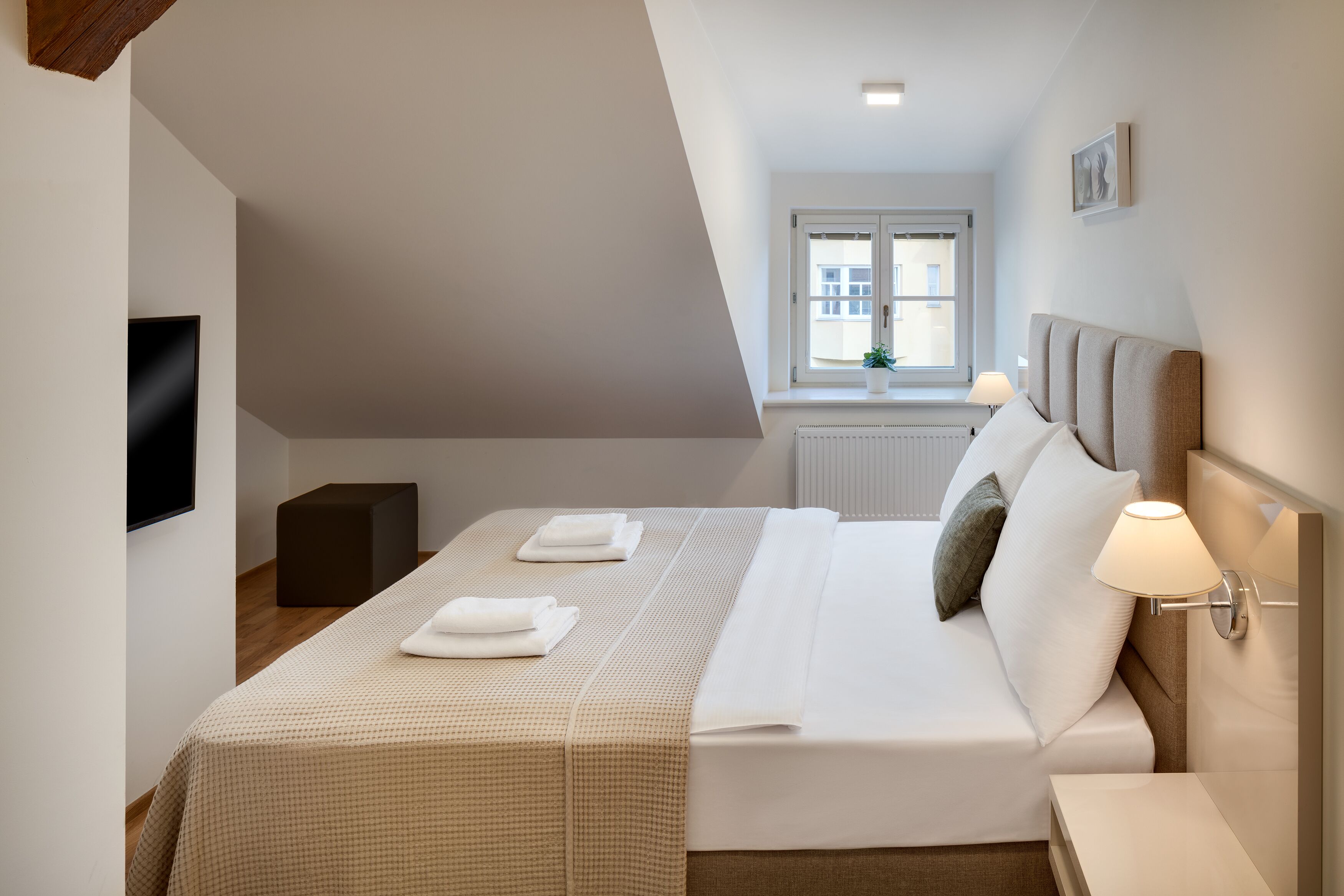 Une chambre moderne avec un lit, des lampes de chevet, un téléviseur à écran plat et une fenêtre donnant sur des immeubles.