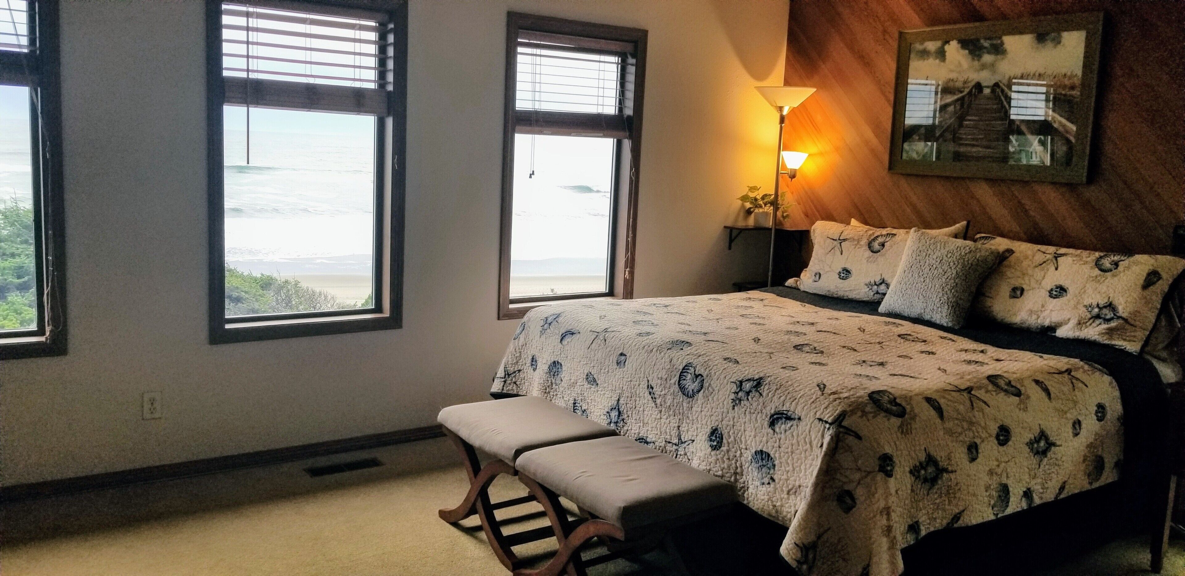 A bedroom with a bed, bench, two windows, a lamp, and a painting on the wall.