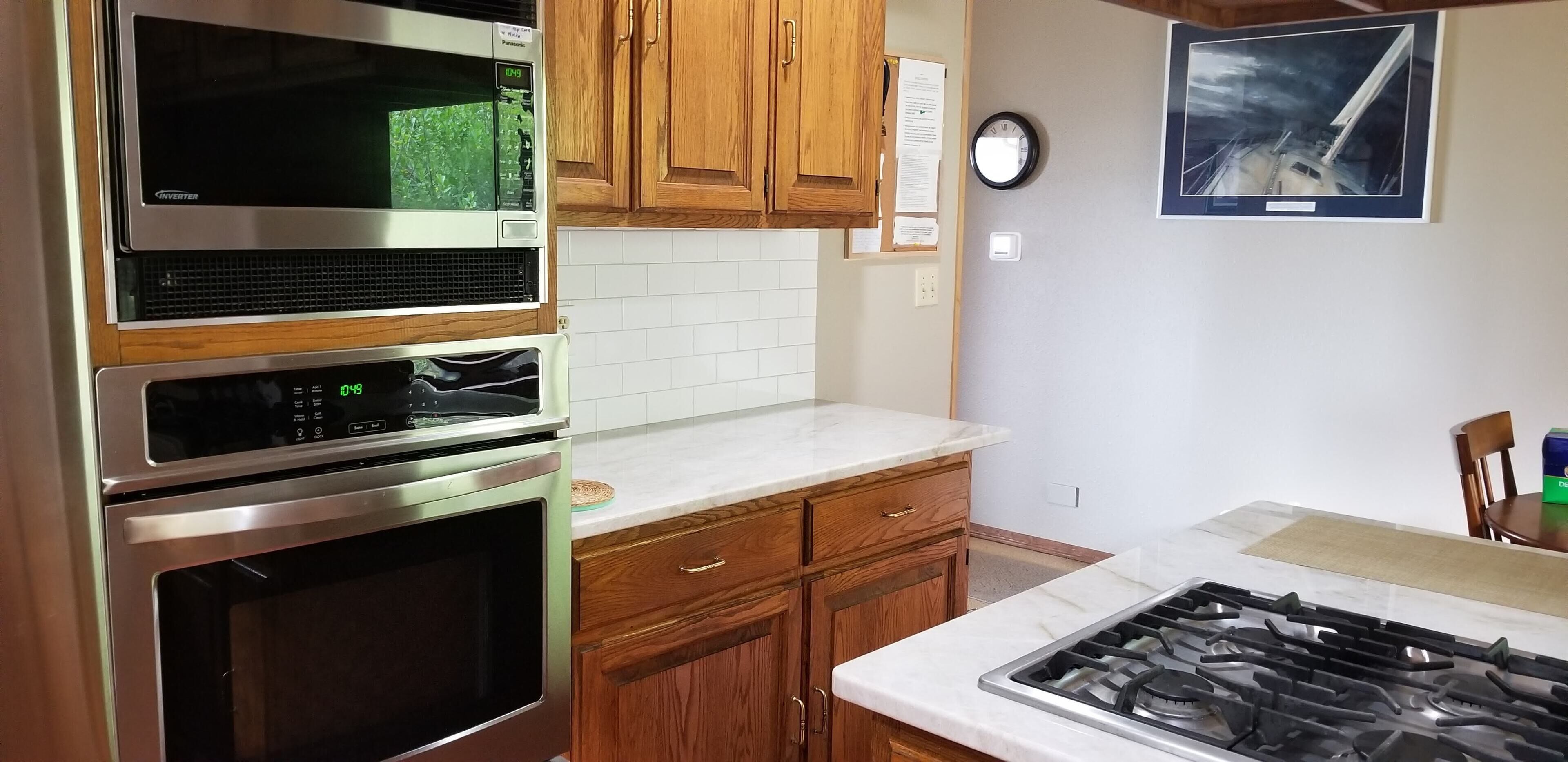 A kitchen with a stainless steel range, a microwave, and a built-in oven. There are wooden cabinets, a white tiled backsplash, and a clock on the wall.