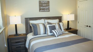 A neatly made bed with striped bedding and multiple pillows, two bedside lamps, a wooden headboard, a small bedside table with a clock, and a motivational quote on the wall. - Guadalupe Bluffs RG 304 Apartment 3 New (New Braunfels)