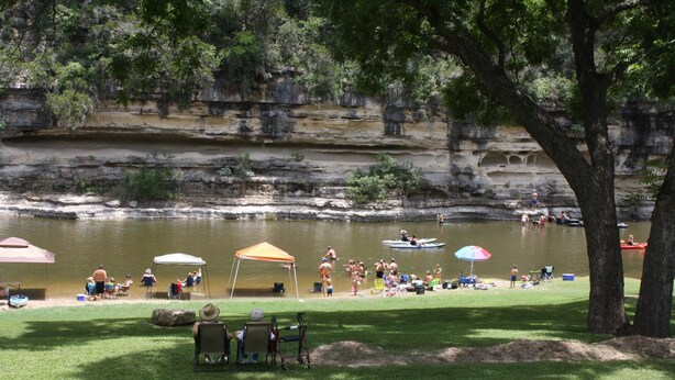 A lakeside park with grassy areas, picnic tables, and a rocky cliff in the background. - Guadalupe Bluffs RG 304 Apartment 3 New (New Braunfels)