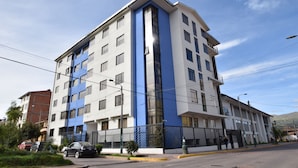 Un edificio de varios pisos con fachada blanca y detalles azules, que cuenta con balcones y una entrada protegida por una reja.