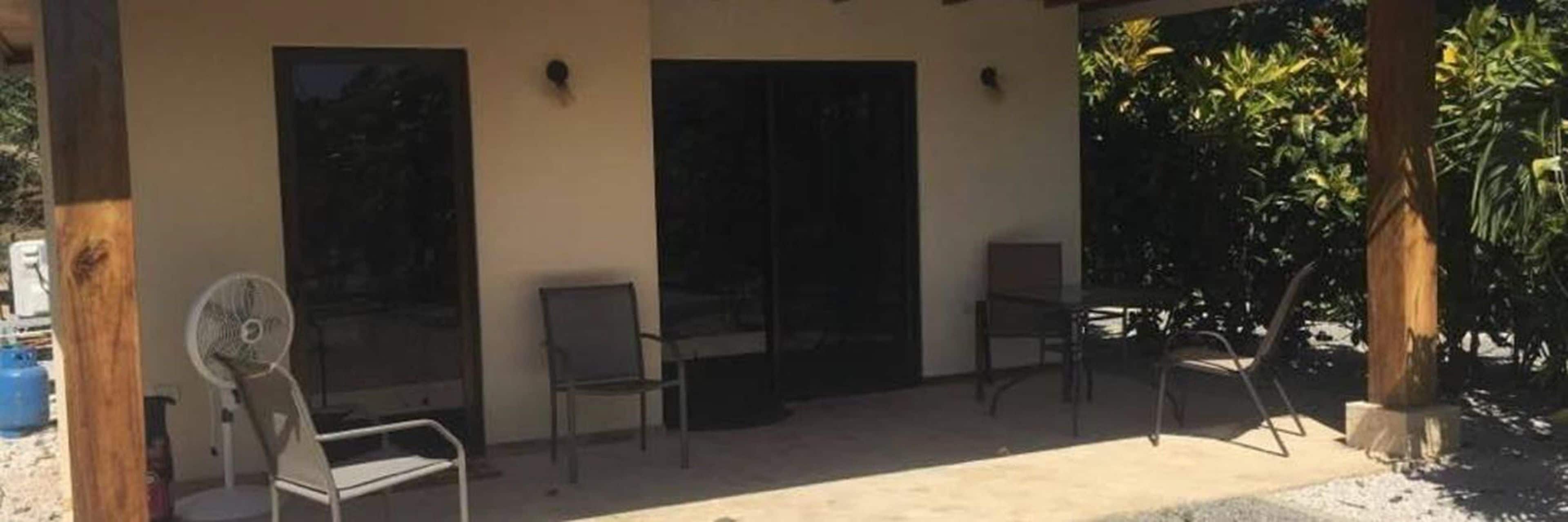 Un espacio al aire libre techado con dos sillas y un ventilador, una puerta y vistas a zonas verdes.