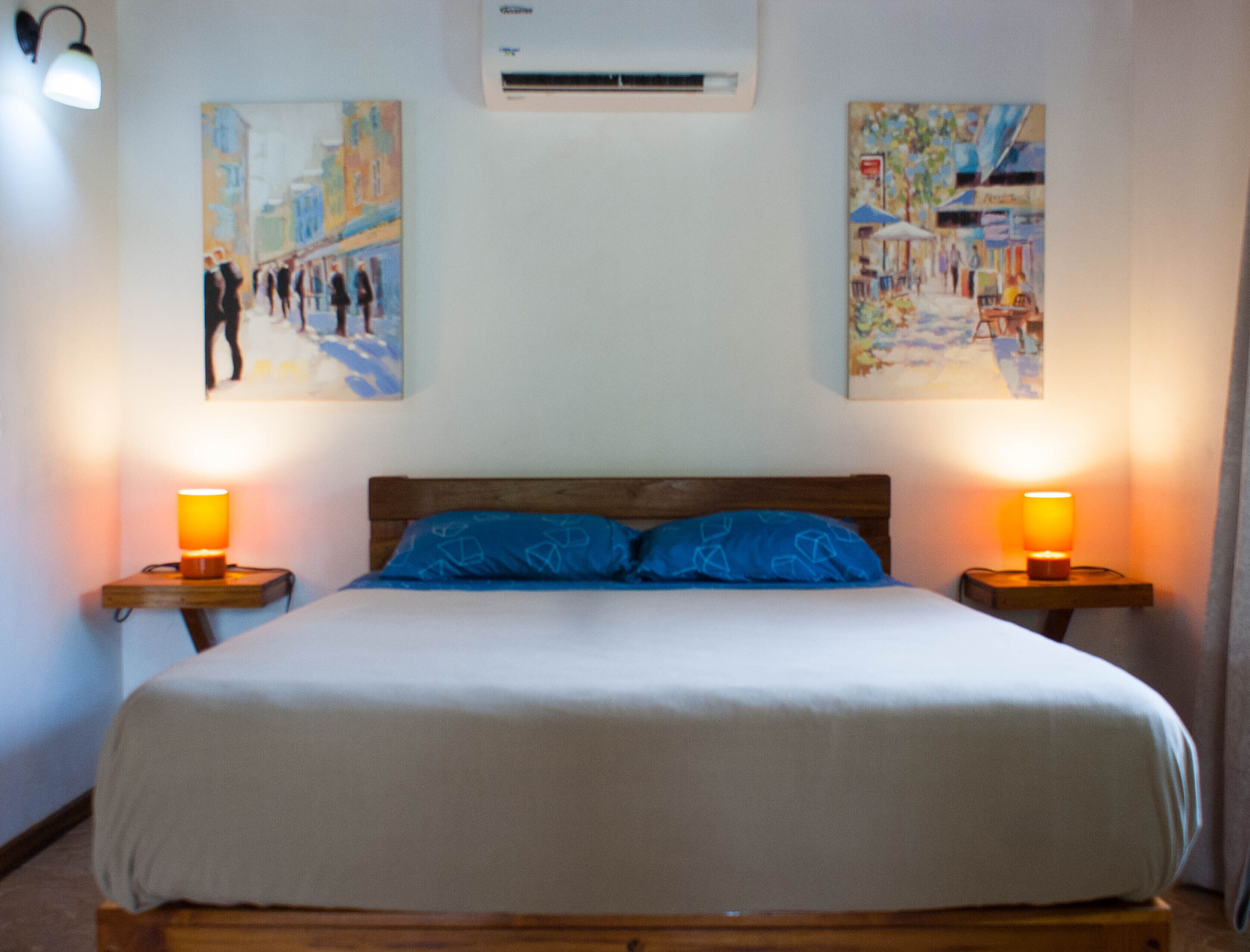 A bedroom with a bed, two bedside lamps, and two paintings on the wall.