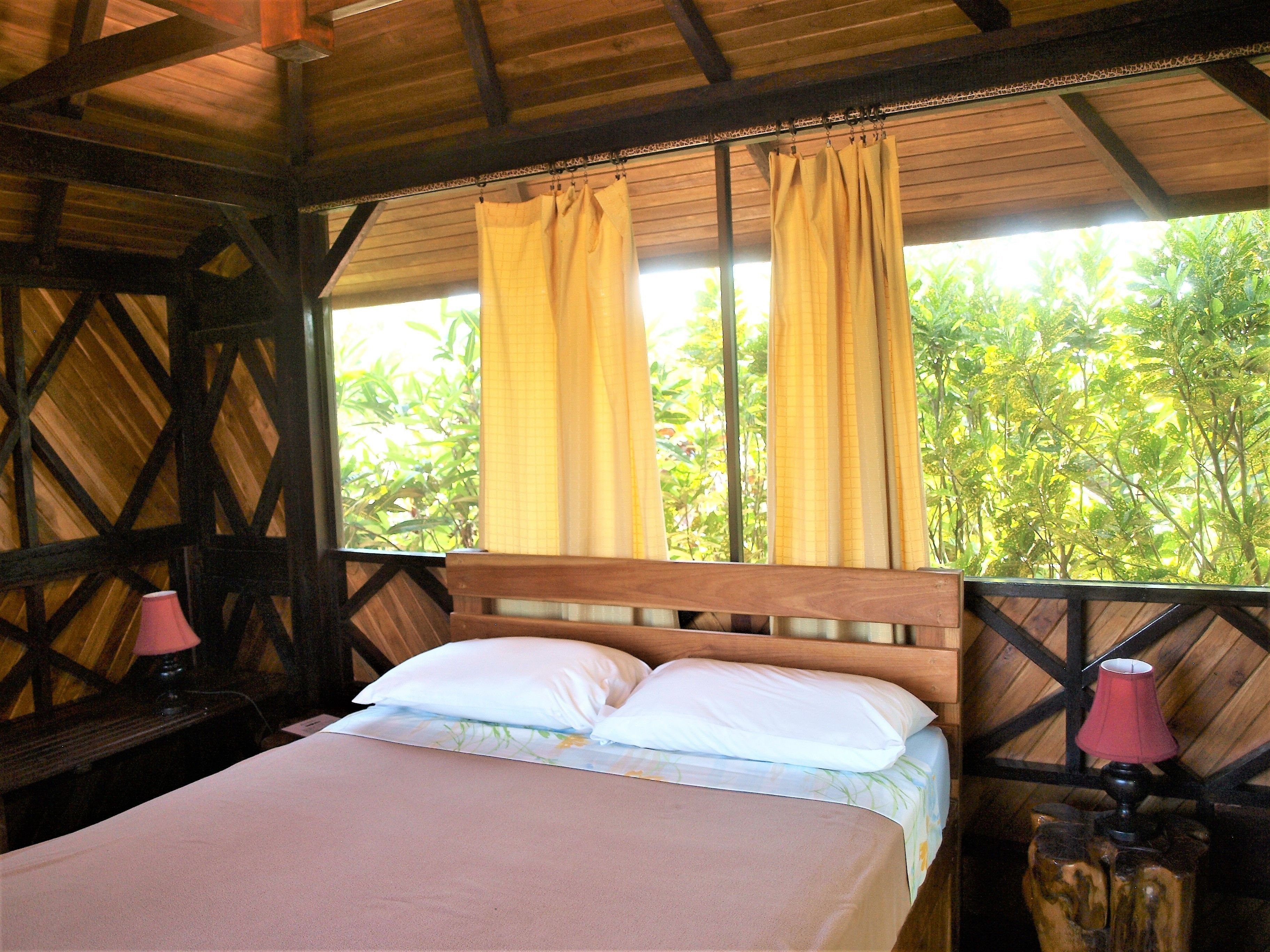 A bed with white linens, a wooden headboard, and a view of greenery through curtains.