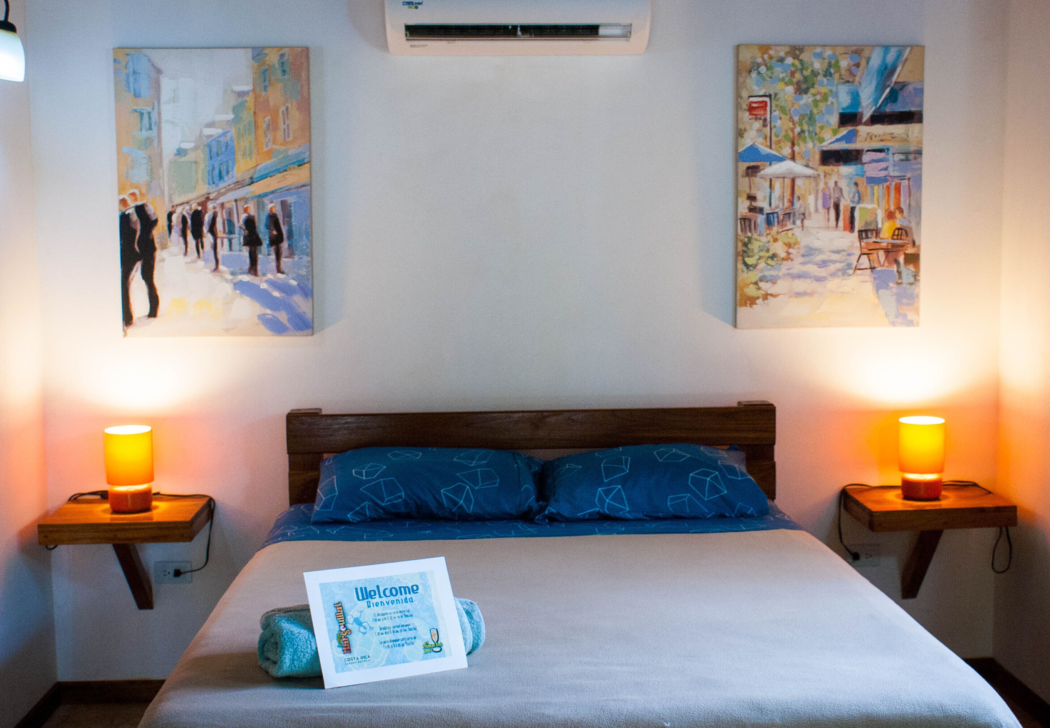 Habitación de hotel con cama, dos lámparas de noche, un cartel de bienvenida y dos cuadros en las paredes.
