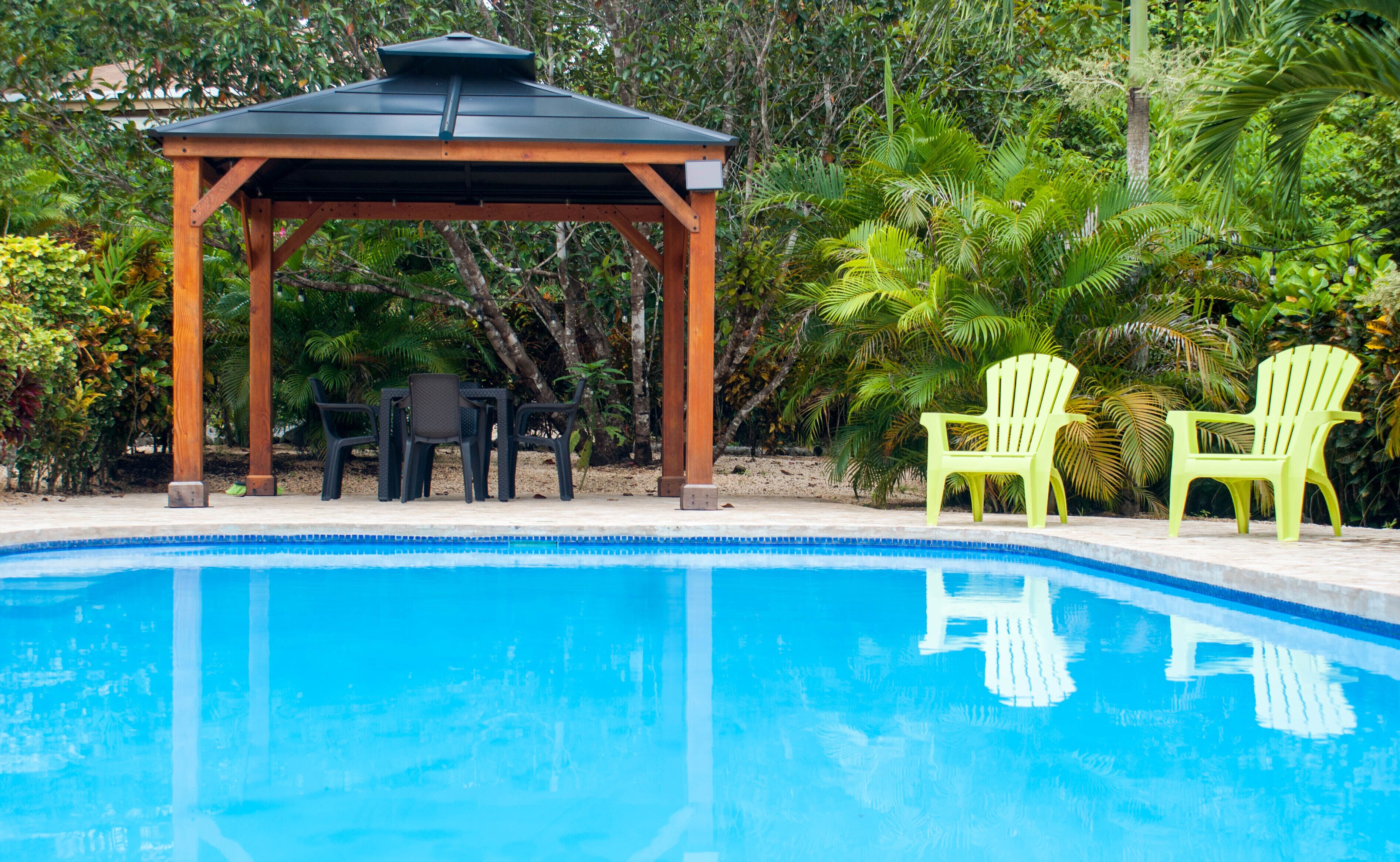Una piscina con un pabellón de madera, dos sillas verdes y una mesa con sillas.