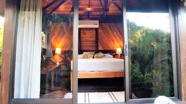 A bedroom with a bed, two bedside lamps, and a view of greenery through large windows. - Lodge Margouillat (Paquera)
