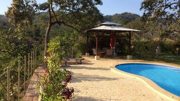 Un área de piscina con muelle de madera, zona de estar cubierta y pérgola.