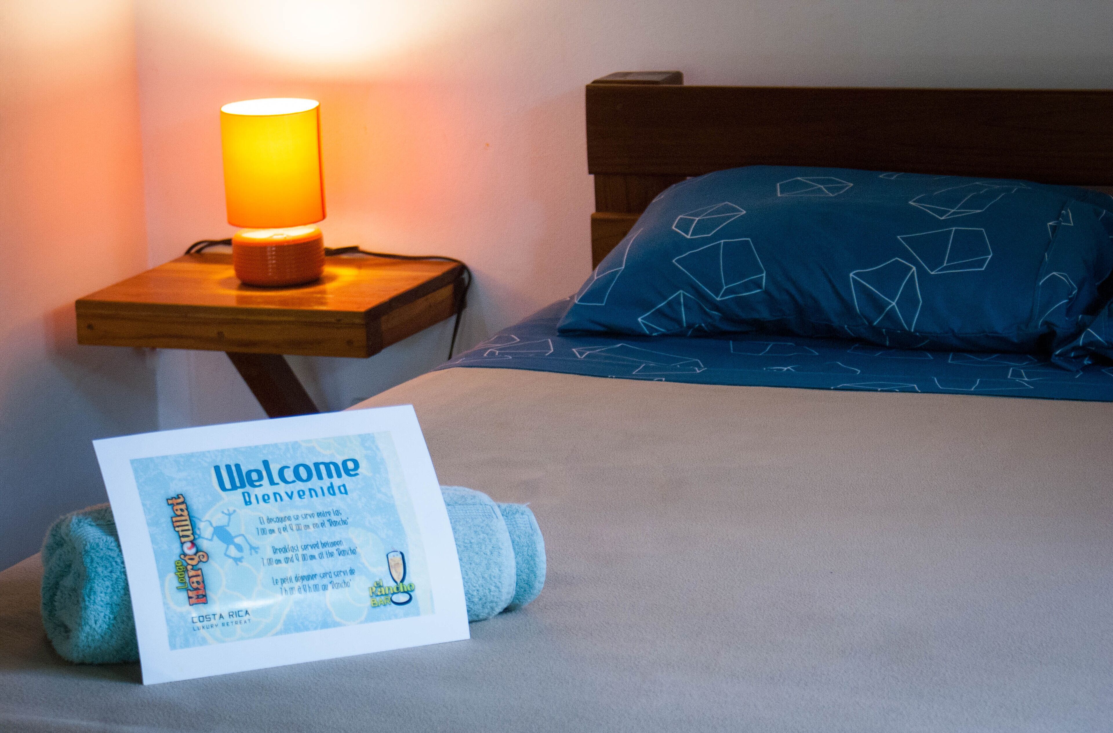 Ein ordentlich bezogenes Bett mit blauem Kissen mit geometrischen Mustern, ein Nachttisch aus Holz mit einer beleuchteten Lampe und eine Willkommenskarte auf dem Bett.