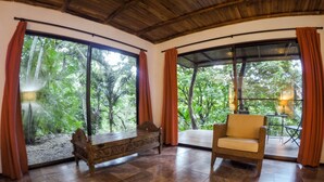 A room with a wooden bench and a cushioned chair, large windows with curtains, and a view of lush greenery. - Lodge Margouillat (Paquera)