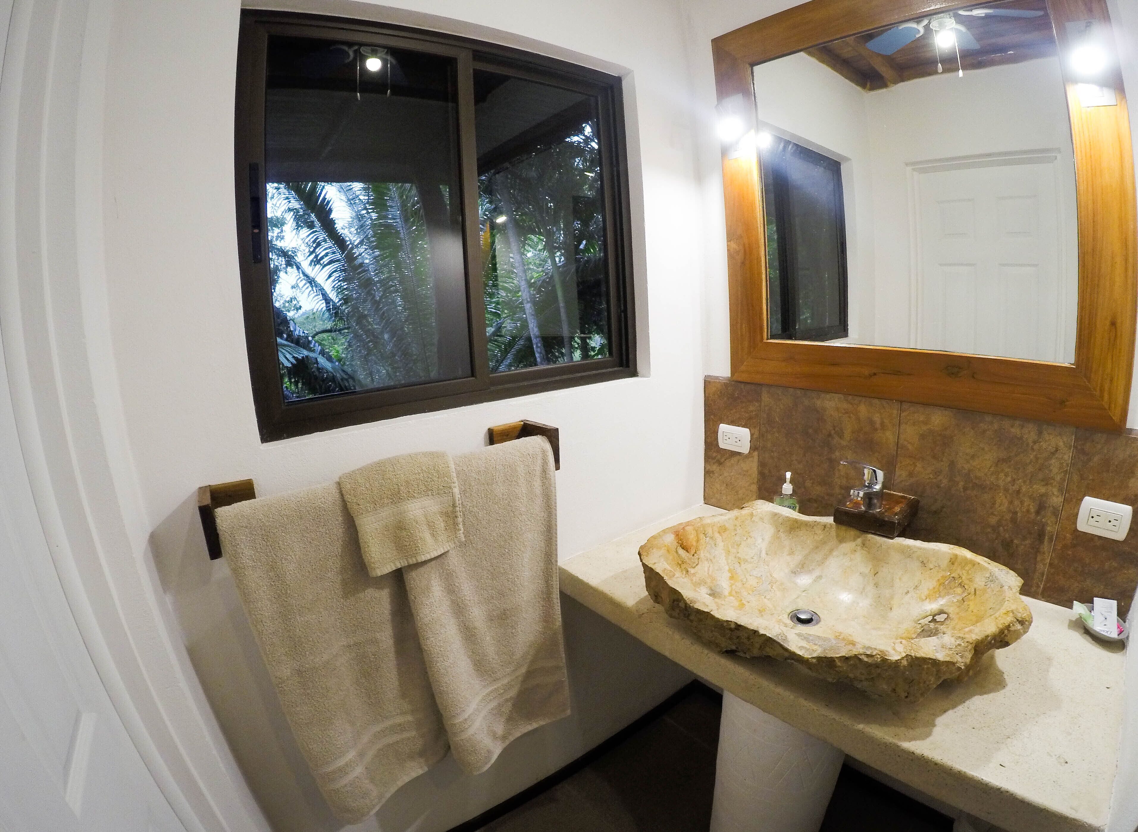 A bathroom with a stone sink, a wooden-framed mirror, and a window with a view of trees.