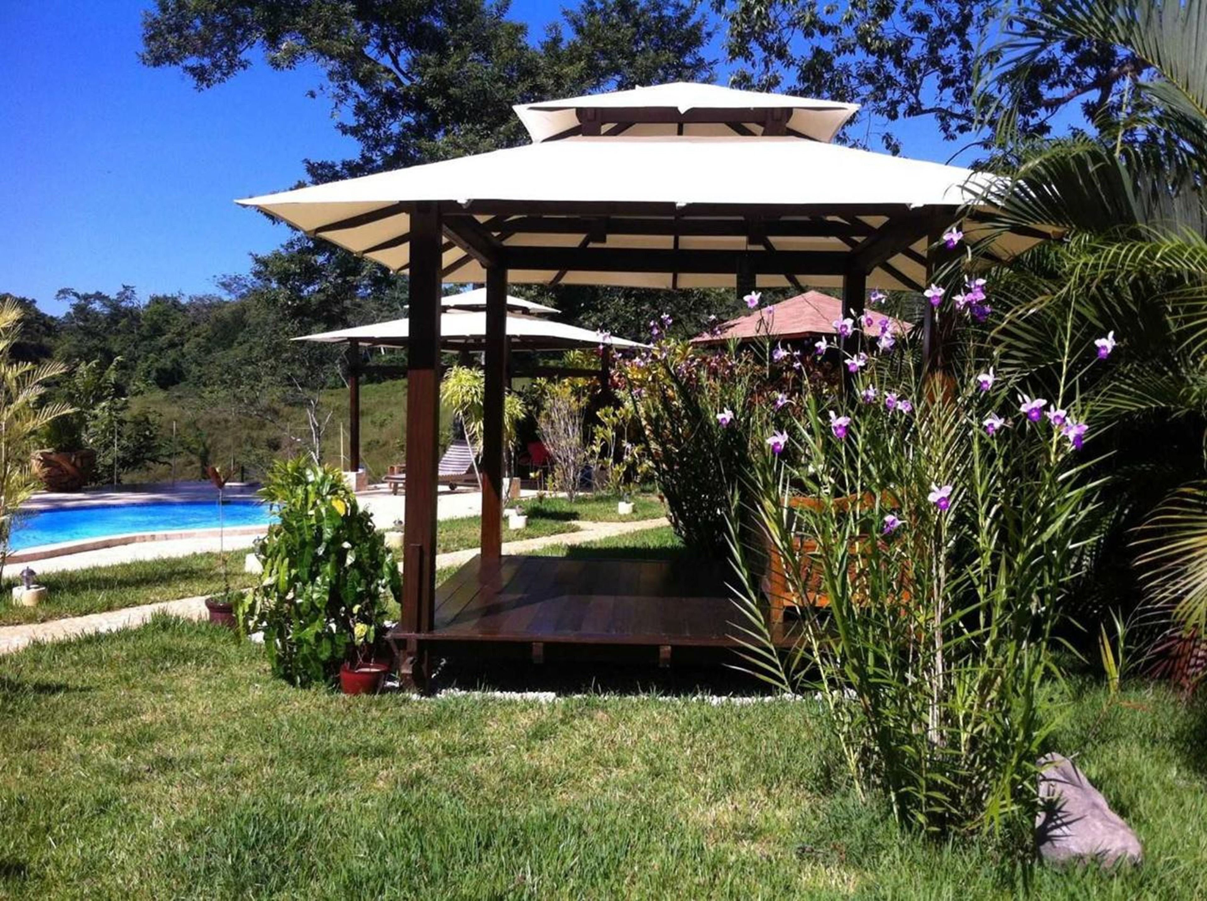 A gazebo with a roof, surrounded by grass and plants, near a swimming pool.