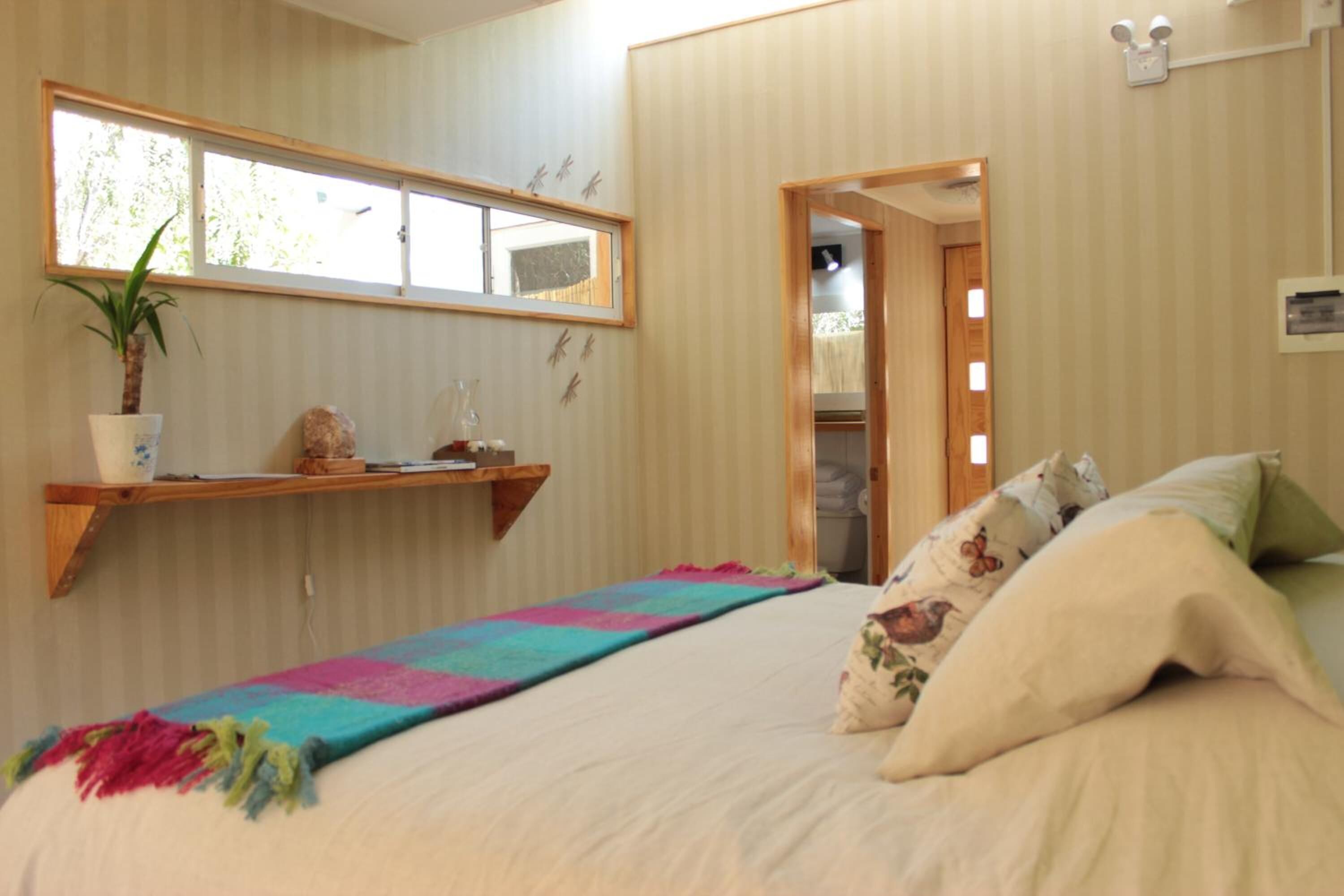 Une chambre à coucher avec un lit, une couverture colorée, une plante en pot, une étagère avec des objets décoratifs et une fenêtre donnant sur un paysage verdoyant.