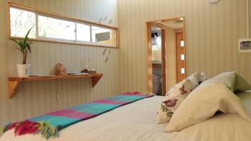 Une chambre à coucher avec un lit, une couverture colorée, une plante en pot, une étagère avec des objets décoratifs et une fenêtre donnant sur un paysage verdoyant.