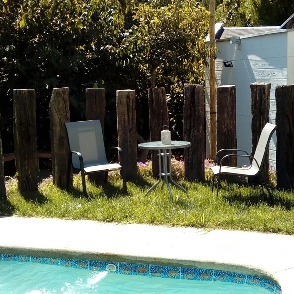 Zona junto a la piscina con una pequeña mesa y dos sillas, rodeada por una valla de madera y un césped.