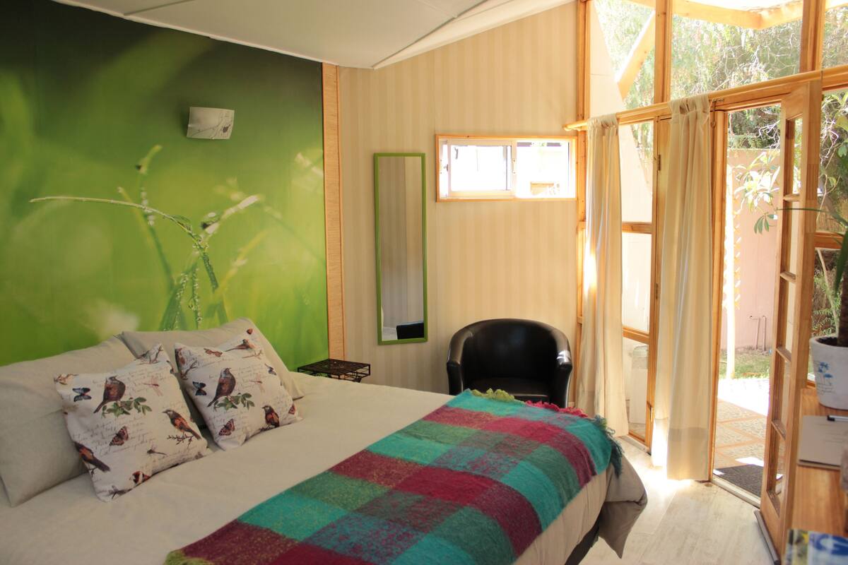Un dormitorio con cama, almohadas, una silla, una ventana y una puerta que da al exterior.