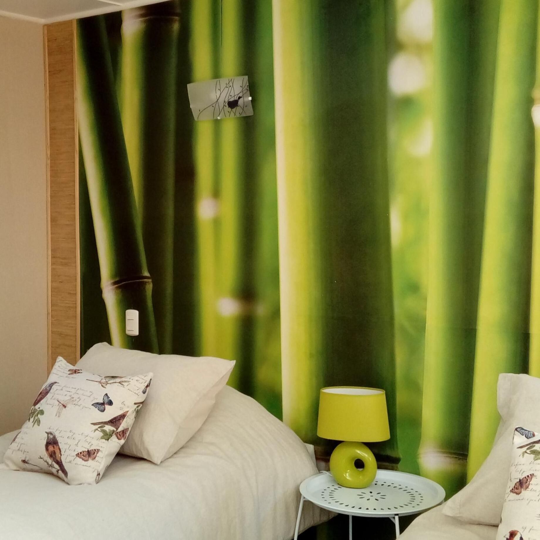 Une chambre d’hôtel avec un lit, des oreillers ornés de motifs d’oiseaux, une petite table ronde avec une lampe jaune et des rideaux verts.