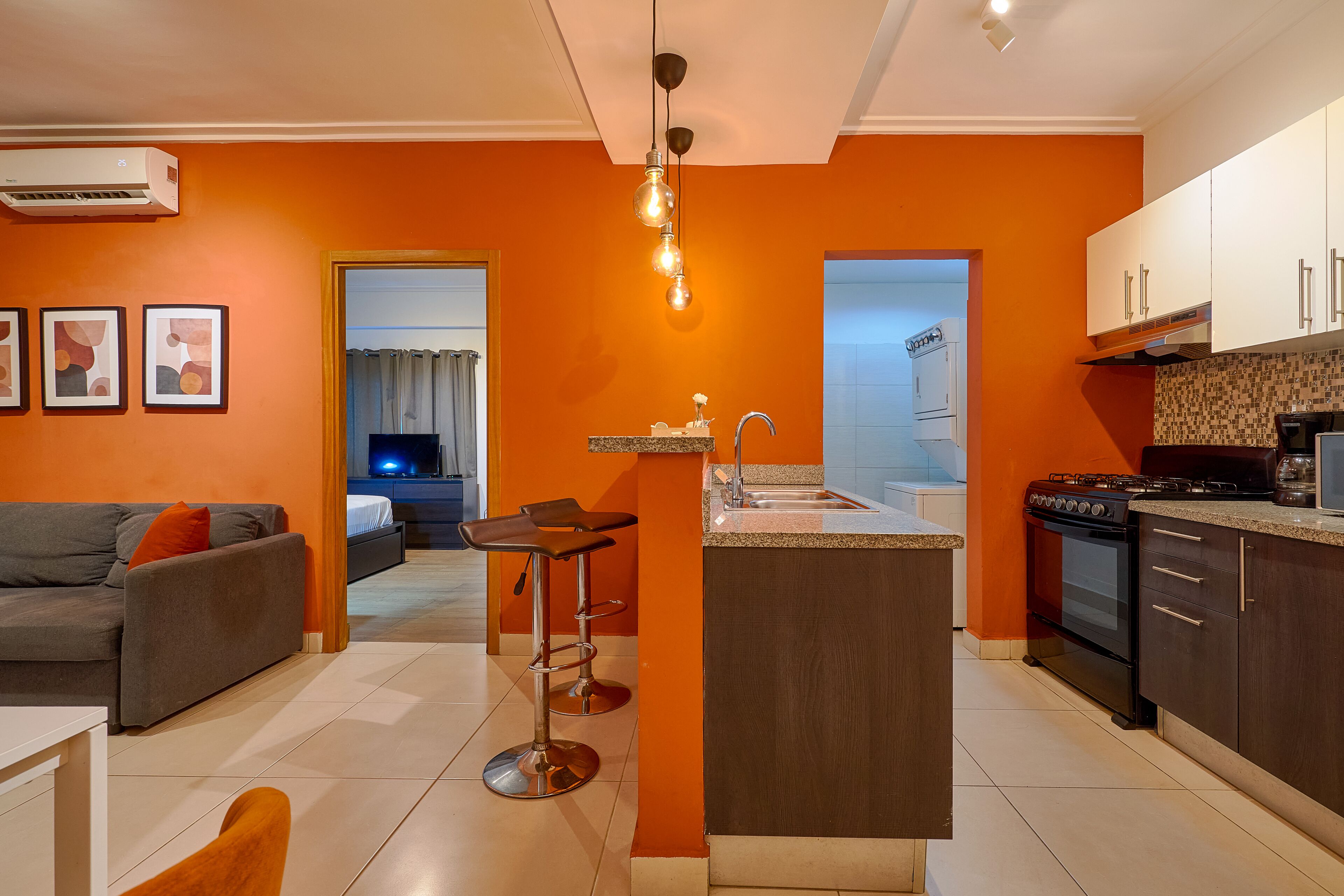 Une cuisine moderne avec un îlot central, un coin repas avec un canapé gris et des coussins orange, et une salle de bain ouverte avec une douche.