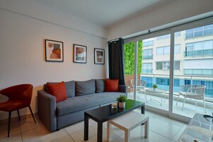 Una sala de estar moderna con un sofá gris, cojines rojos y negros, una mesa de centro negra y un amplio ventanal corredero que da a un balcón con mobiliario exterior.