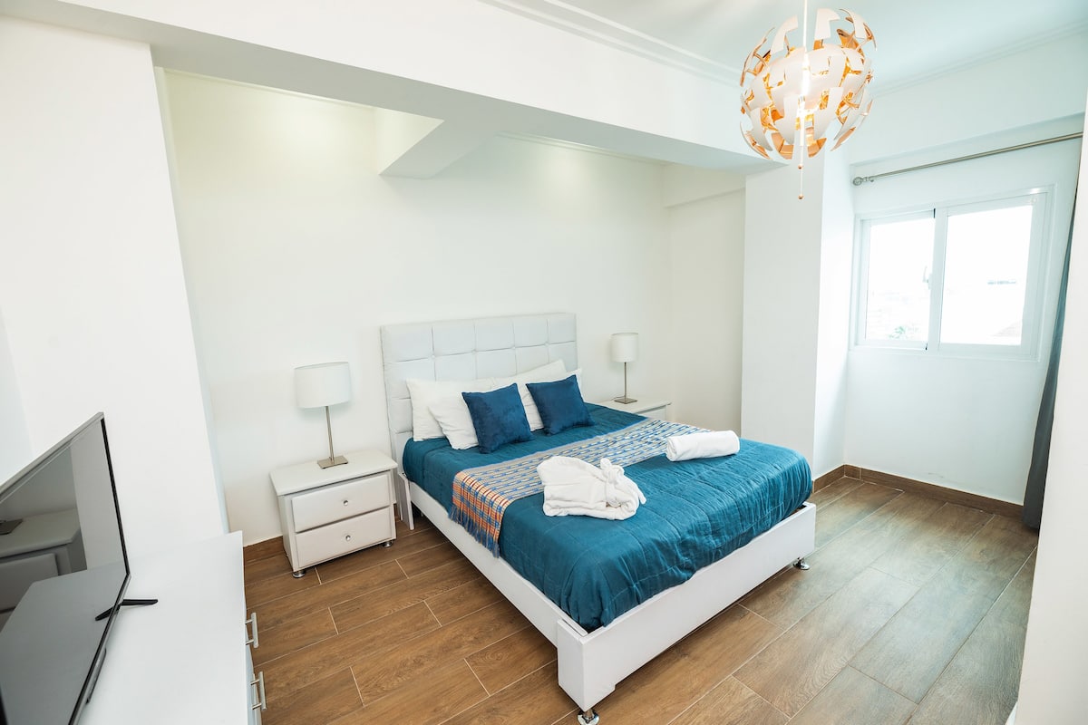 Een moderne slaapkamer met een wit bed, blauw beddengoed en een houten vloer.